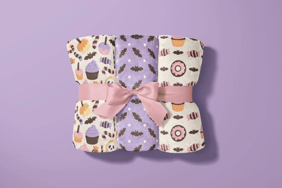 Three Halloween-Themed Blankets Rolled And Tied With A Pink Ribbon.  The Blankets Feature Different Patterns Of Candy, Bats, And Cupcakes.