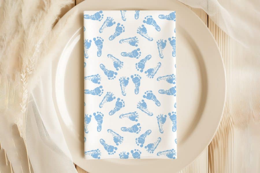 A White Napkin With A Pattern Of Light Blue Baby Footprints On A Beige Plate.