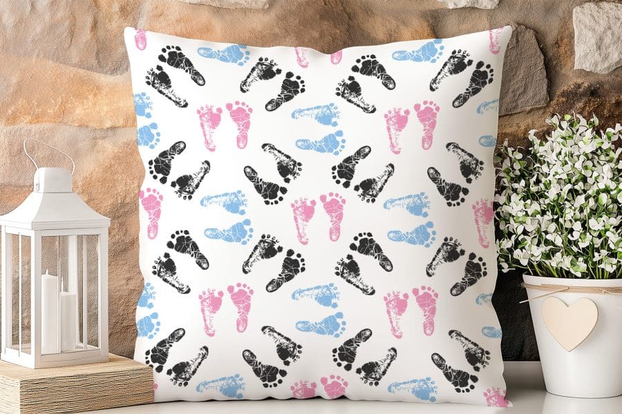 White Square Pillow With A Repeating Pattern Of Black, Pink, And Blue Baby Footprints.