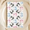 A White Napkin With A Pattern Of Pink, Blue, And Black Baby Footprints Sits On A Beige Plate.