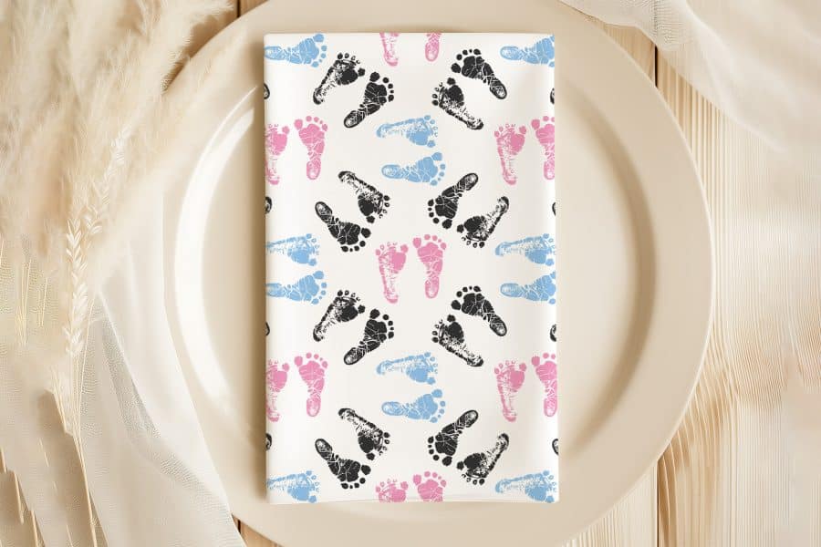 A White Napkin With A Pattern Of Pink, Blue, And Black Baby Footprints Sits On A Beige Plate.
