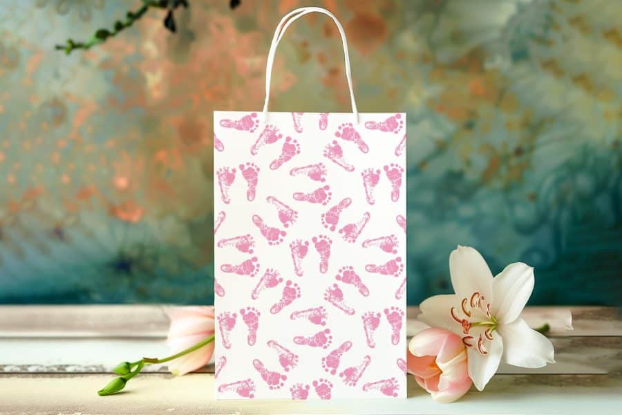 White Gift Bag With Pink Baby Footprints, Next To Pink Flowers.
