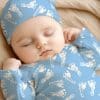 Sleeping Baby Wearing A Light Blue Footed Sleeper And Hat With A White Footprint Pattern.