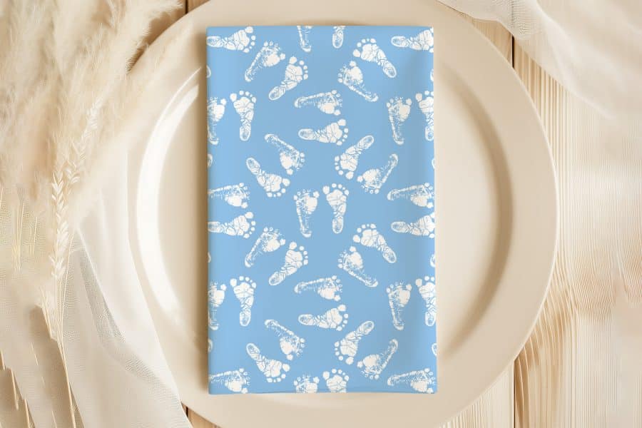 Light Blue Napkin With A Pattern Of White Baby Footprints, Placed On A Beige Plate.