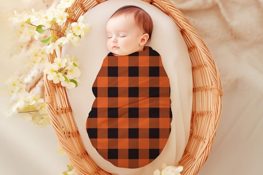 Sleeping baby swaddled in an orange and black buffalo plaid blanket.