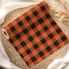 A Roll Of Orange And Black Buffalo Plaid Fabric Rests On A Woven Mat.