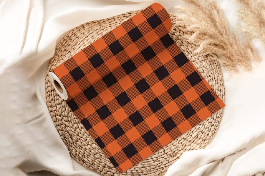 A Roll Of Orange And Black Buffalo Plaid Fabric Rests On A Woven Mat.
