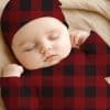 Sleeping Baby Wearing A Red And Black Buffalo Plaid Onesie And Matching Hat.