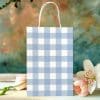 A Light Blue And White Gingham Gift Bag Sits On A Wooden Surface Next To Pink Tulips And A White Lily.