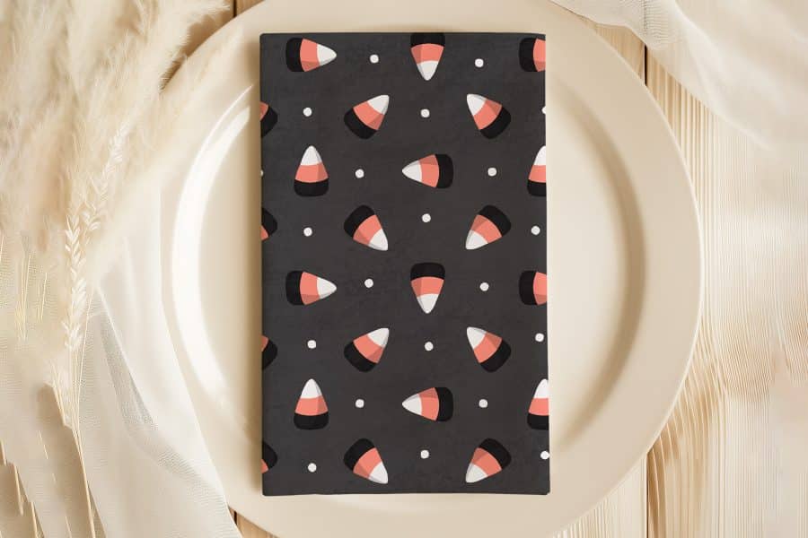 A Dark Gray Napkin With A Repeating Pattern Of Candy Corn Sits On A Beige Plate.