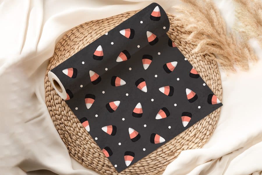 A Roll Of Dark Gray Fabric With A Repeating Pattern Of Candy Corn Sits On A Woven Placemat.