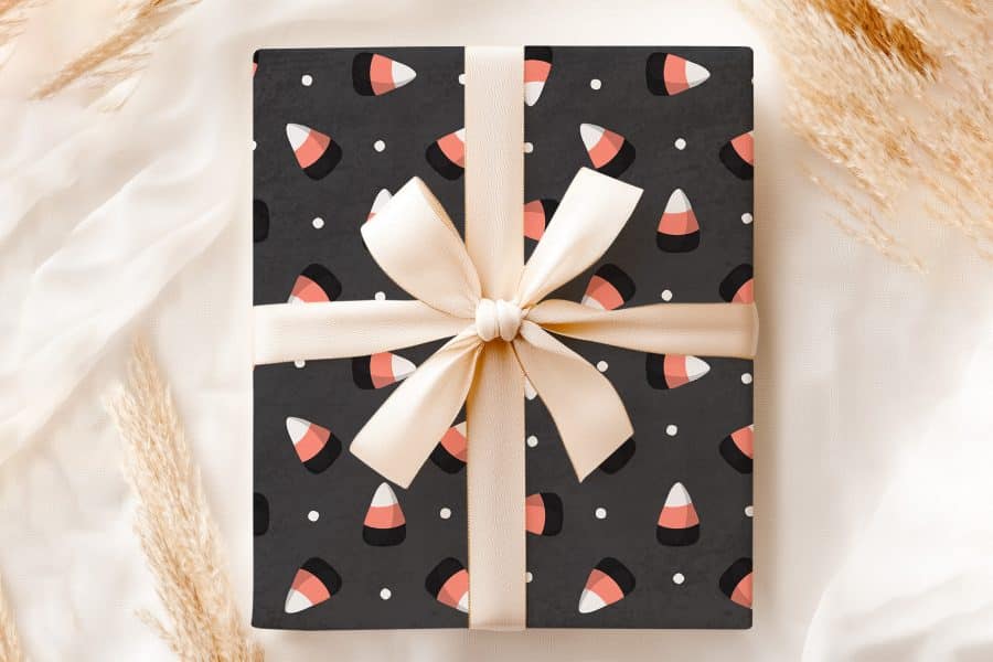 A Gift Wrapped In Dark Gray Paper Featuring A Repeating Pattern Of Candy Corn, Tied With A Beige Ribbon.