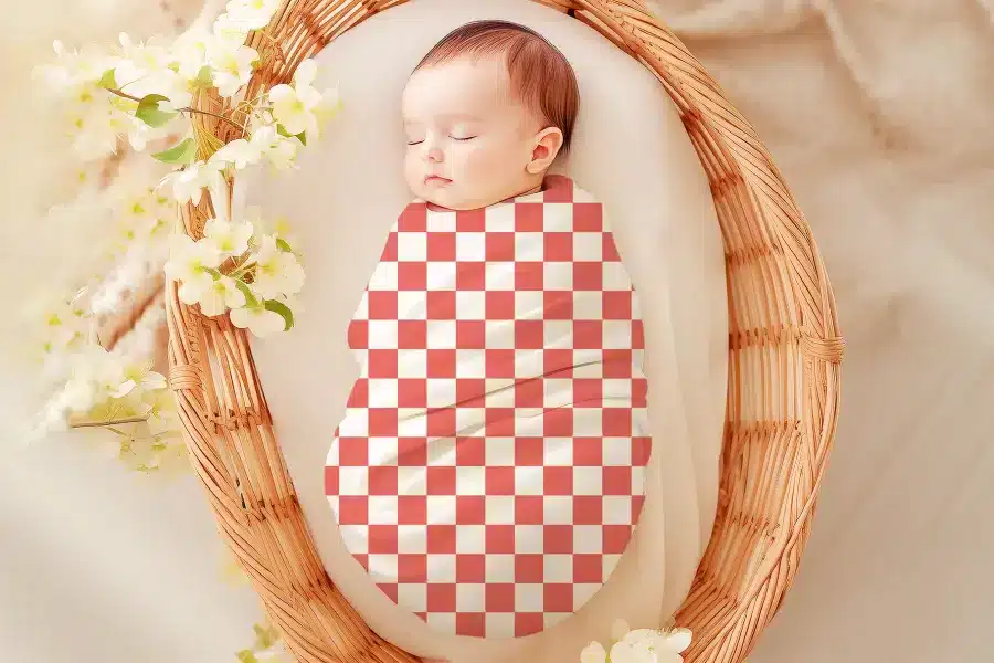 Sleeping baby swaddled in a coral and white checkered receiving blanket.