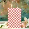 A Coral And White Checkered Paper Gift Bag Sits On A Table In Front Of Blurred Flowers.