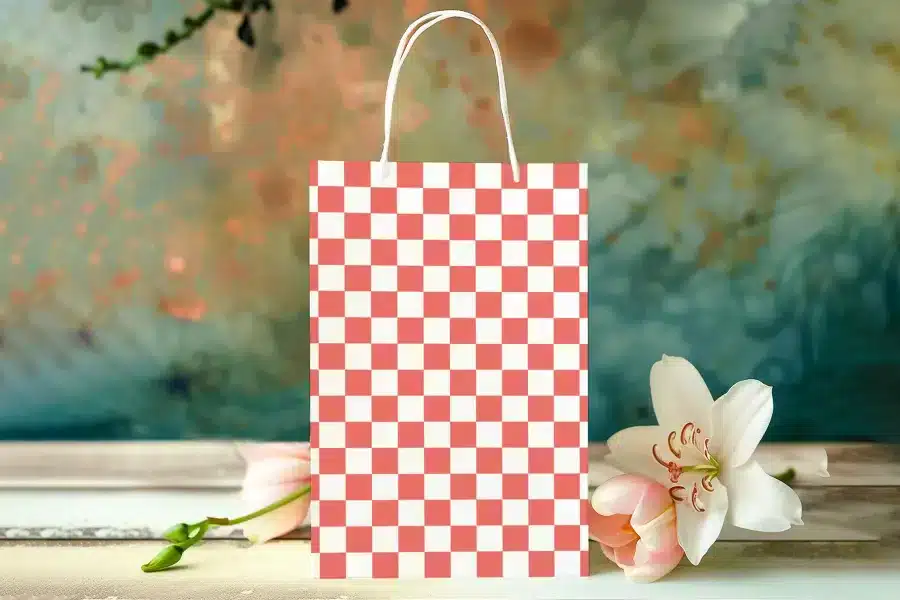A Coral And White Checkered Paper Gift Bag Sits On A Table In Front Of Blurred Flowers.
