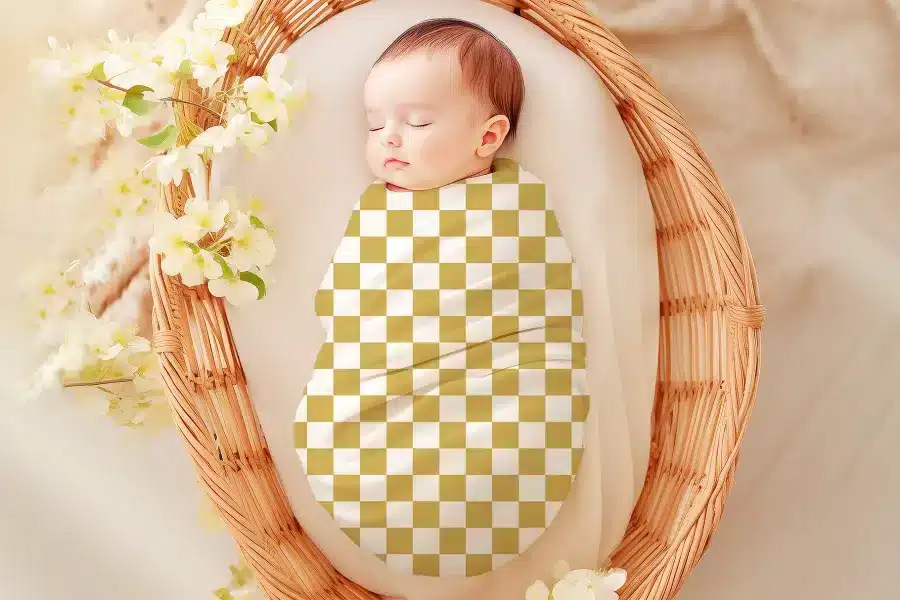 Sleeping baby swaddled in a gold and white checkered blanket inside a wicker basket.