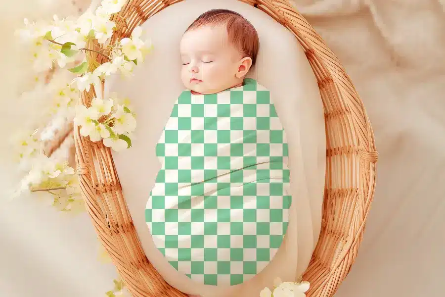 Sleeping baby swaddled in a mint green and white checkered wrap, nestled in a wicker basket.
