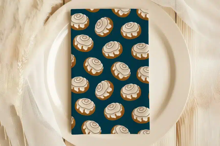A Dark Teal Napkin With A Repeating Pattern Of Cinnamon Rolls Sits On A White Plate.