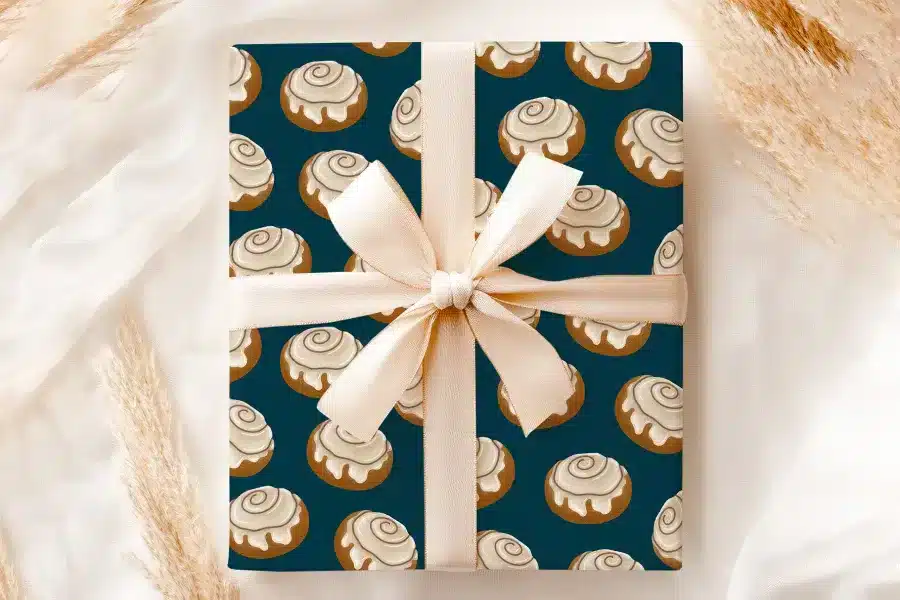 A Gift Wrapped In Teal Paper With A Repeating Pattern Of Cinnamon Rolls, Tied With A Beige Ribbon.
