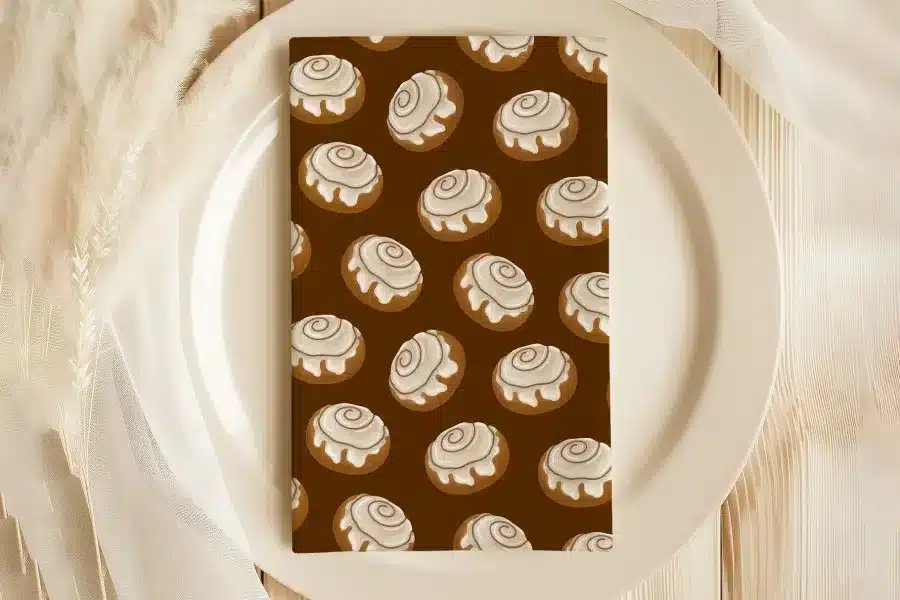 A Brown Napkin With A Repeating Pattern Of Cinnamon Rolls Sits On A White Plate.