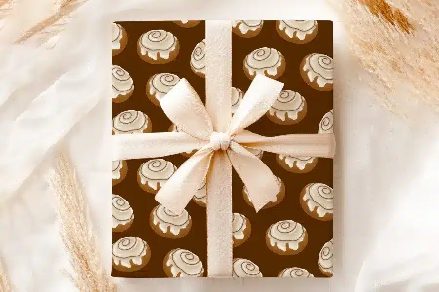 A Gift Wrapped In Brown Paper Featuring A Repeating Pattern Of Cinnamon Rolls, Tied With A Beige Ribbon.