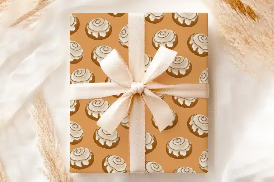 Gift Wrapped In Beige Paper With A Pattern Of Cinnamon Rolls And Tied With A Cream-Colored Ribbon.