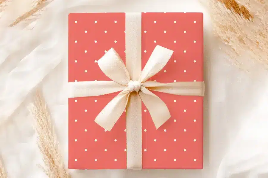 A Coral-Colored Gift Box With White Polka Dots, Tied With A Cream-Colored Ribbon And Bow.