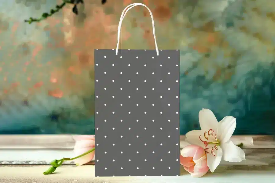 Gray Gift Bag With White Polka Dots, Sitting On A Table With Pink And White Flowers.