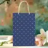 Navy Blue Gift Bag With White Polka Dots, Sitting On A Table With Pink Flowers.