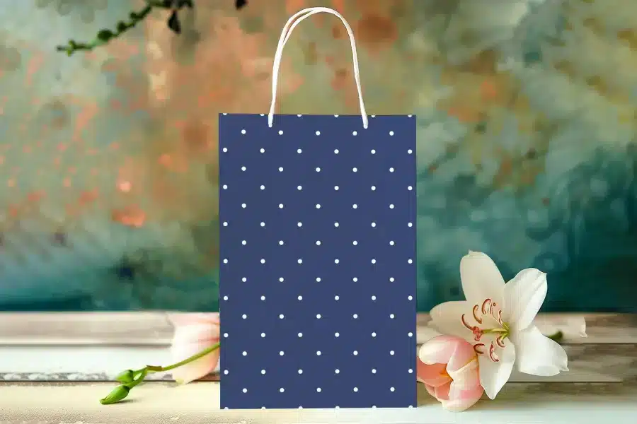 Navy Blue Gift Bag With White Polka Dots, Sitting On A Table With Pink Flowers.