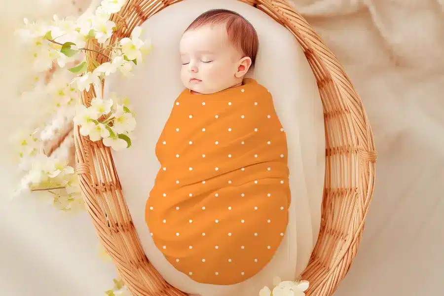 Sleeping baby swaddled in an orange polka dot wrap inside a wicker basket.