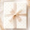 A Gift, Wrapped In Off-White Paper With Gold Polka Dots And Tied With A Beige Ribbon.