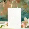 White Gift Bag With Light Pink Polka Dots, Flanked By Pink Tulips And A White Lily.