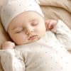 A Sleeping Baby Wearing A White Polka Dot Onesie And Matching Hat.