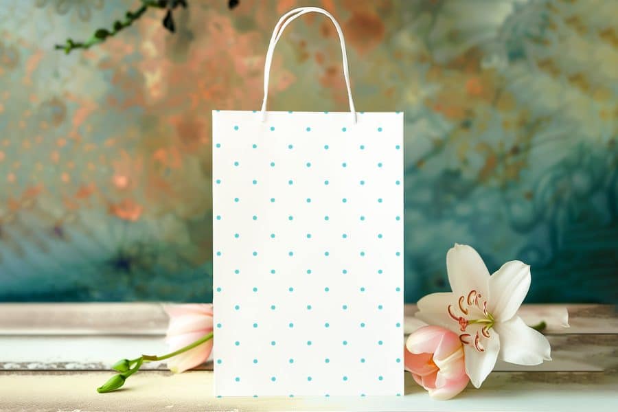 White Paper Gift Bag With Light Teal Polka Dots, Situated In Front Of Blurred Flowers.