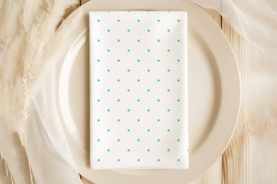 White Dinner Napkin With Light Teal Polka Dots On A Beige Plate.