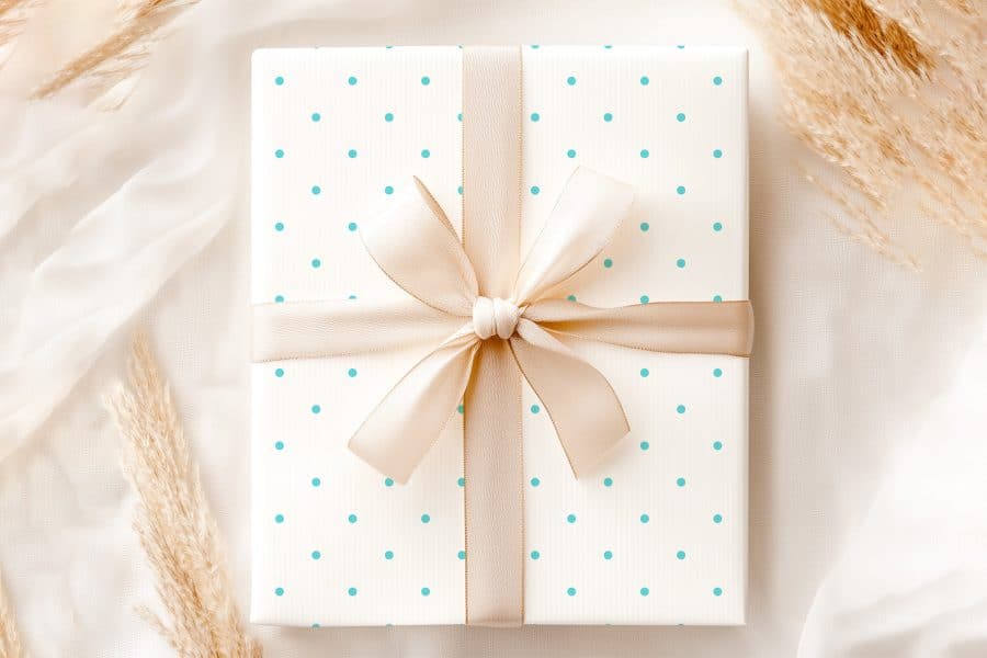 A Gift Box With A Light Beige Ribbon And Light Teal Polka Dots On A White Background.