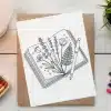 Line Drawing Of Flowers And A Fern Pressed Inside An Open Book, With A Pen Resting On Top.