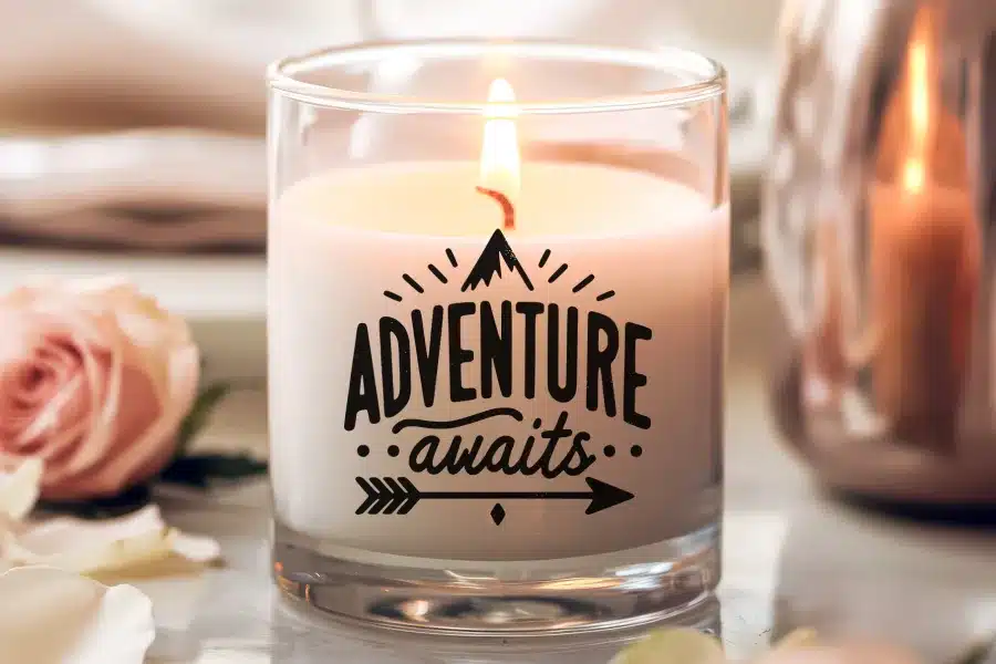 A lit candle in a clear glass jar with the words Adventure awaits printed on the side.