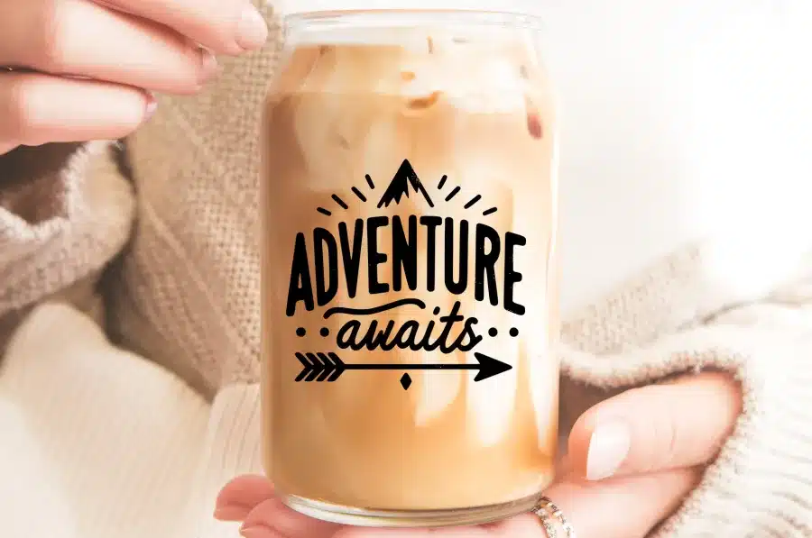 A Person'S Hands Holding A Glass Iced Coffee With The Words Adventure Awaits Printed On It.