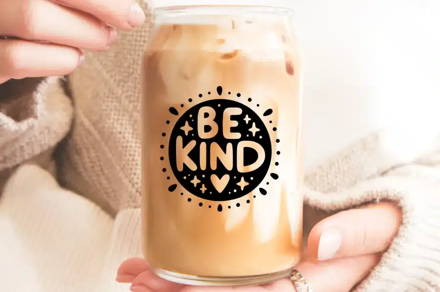 A Person'S Hands Holding A Glass Iced Coffee Can With The Words Be Kind In A Circular Design.