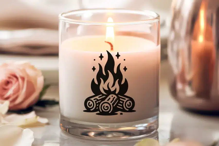 A lit candle in a clear glass jar with a campfire design on the side.