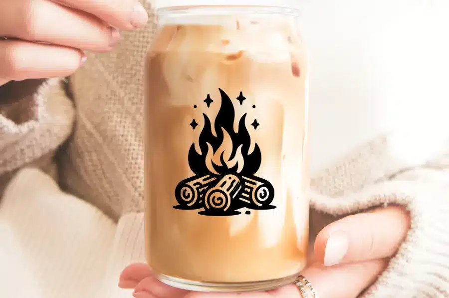 A Person Holds A Glass Iced Coffee With A Campfire Design.