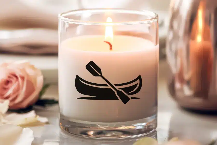 A lit candle in a clear glass jar features a canoe and paddle design.