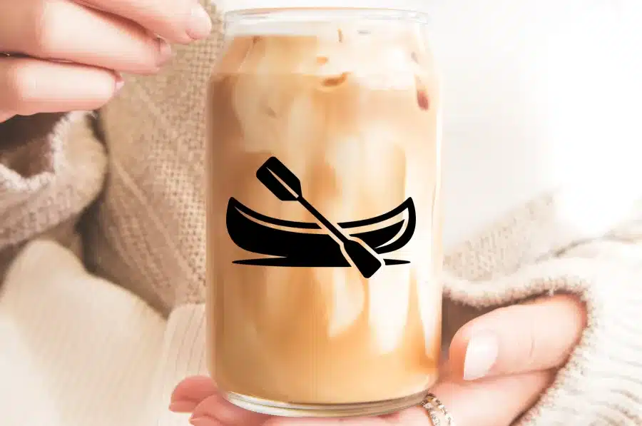 Hands Holding A Glass Jar Filled With Iced Coffee, Featuring A Canoe And Paddle Graphic.