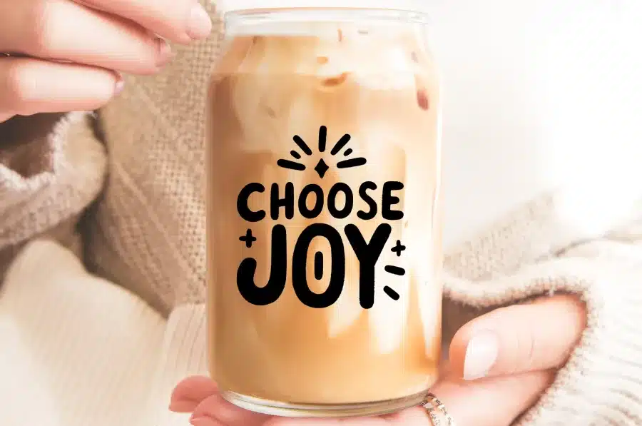 Close-Up Of Hands Holding A Glass Jar Filled With Iced Coffee. The Jar Is Decorated With The Text Choose Joy.