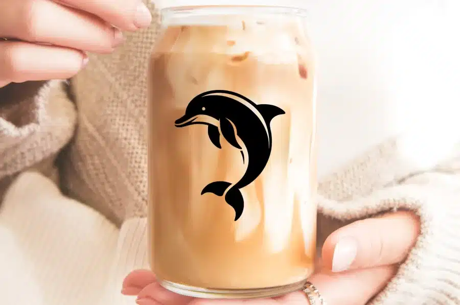 A Person'S Hands Holding A Glass Jar Filled With Iced Coffee, Featuring A Black Dolphin Silhouette On The Side.