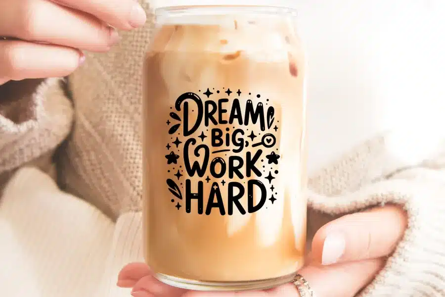 A person's hands holding a glass iced coffee can with the text Dream Big Work Hard.
