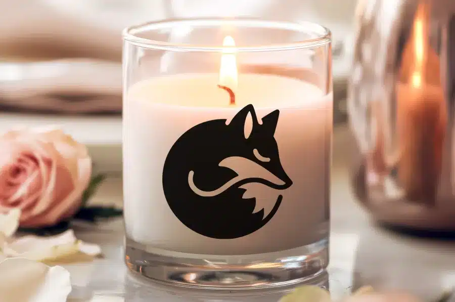 A Lit Candle In A Clear Glass Jar, Featuring A Black Silhouette Of A Curled-Up Fox.