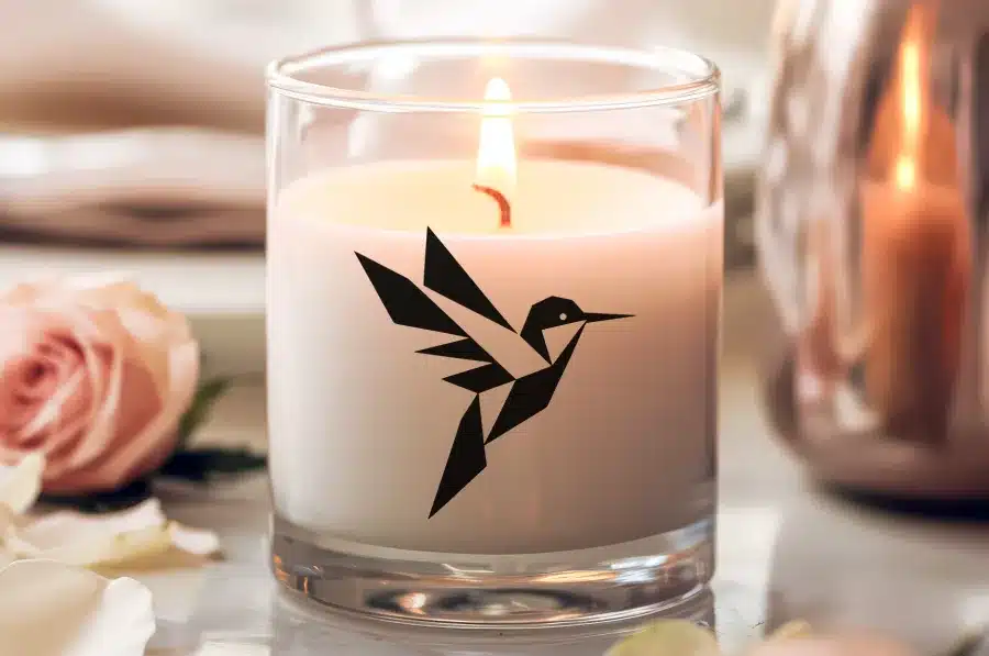 A Lit Candle In A Clear Glass Jar, Featuring A Geometric Hummingbird Design.